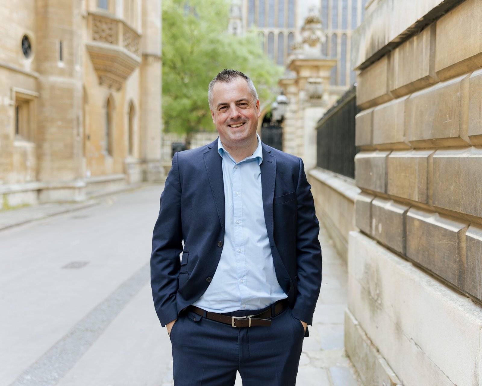 A man in a navy suit and light blue shirt stands with his hands in his pockets on a paved street lined with carved stone buildings.