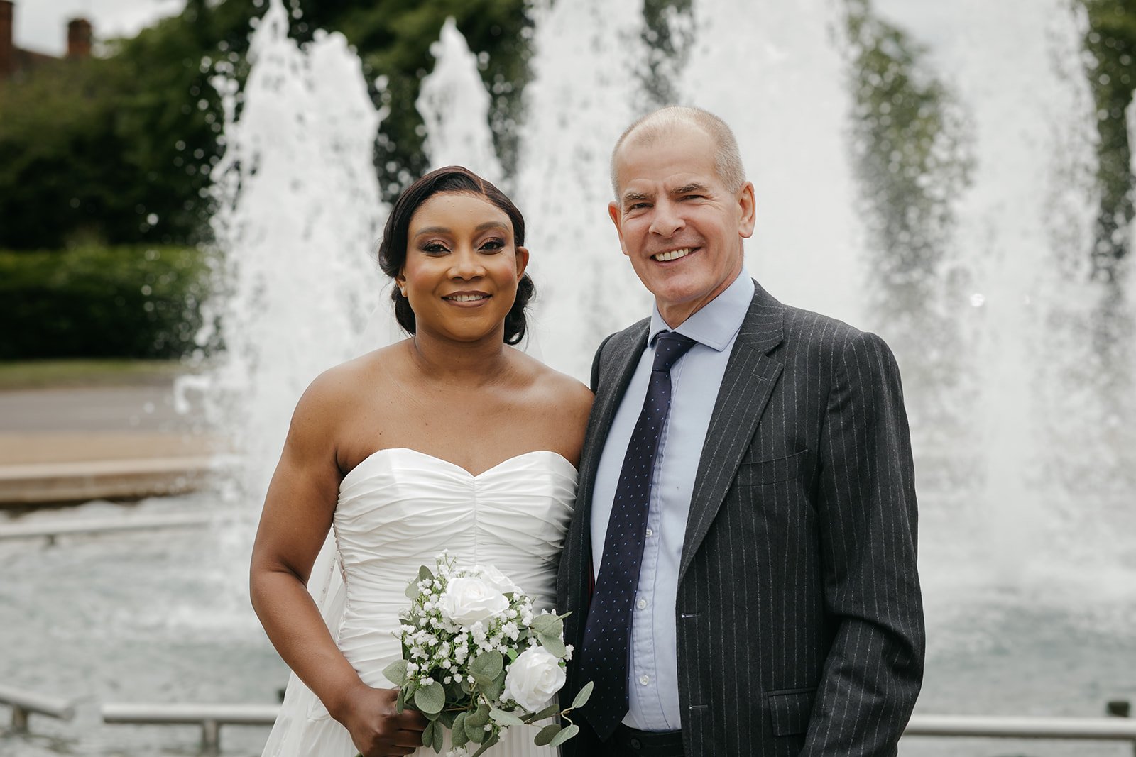 A bride in a strapless white gown holding white roses stands beside a man in a pinstriped grey suit in front of tall jets of a fountain.